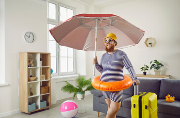 Plus size man in striped swimsuit, hat, shades and floaty standing in living room, holding beach umbrella and luggage and making funny face. Covid 19 pandemic, summer holiday, vacation at home concept - obrazy, fototapety, plakaty