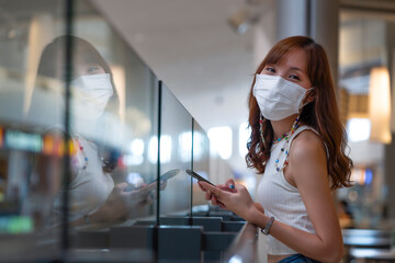 Young attractive Asian woman wear white mask using smartphone at shopping mall with happiness face, use technology to connect website for ordering goods online. Modern shopping online during Covid-19