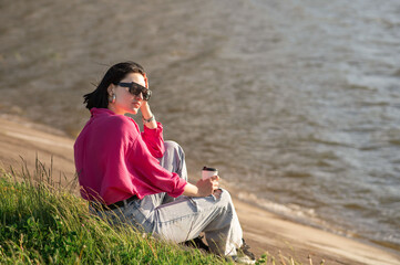 Brunette woman in sunglasses sitting on lakeshore and chilling in the sun rays outdoor