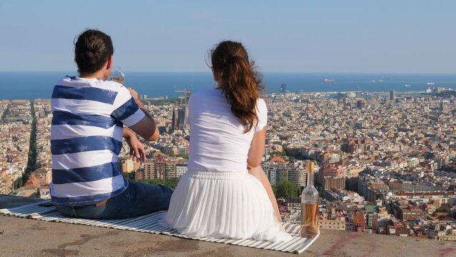 Couple rest at high point, drink wine and enjoy aerial view of Barcelona city at summer evening. Man and woman spend time together, sitting against impressive cityscape