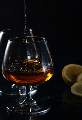 Naklejka na ściany i meble Two cognac glasses on a dark background. Cognac is poured into one glass from above