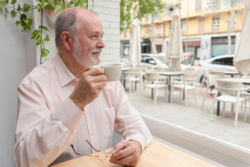 portrait of a smiling retired man looking out the window in a coffee bar holding his cup of coffee and eyeglasses