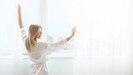 Well-rested blonde lady opening curtains at home - obrazy, fototapety, plakaty