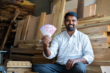 Happy young indian carpenter holding money or Indian currency notes by loooking at camera - concept...
