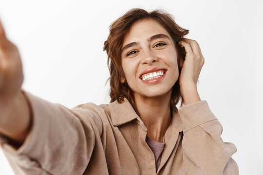 Close up portrait of smiling brunette girl taking selfie, touching her face, posing for photo, picture of herself on mobile phone, standing over white background