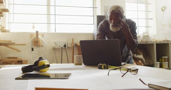 African american male carpenter talking on smartphone and using laptop in a carpentry shop
