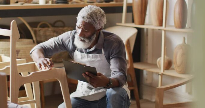 African american male carpenter touching wooden chair frame and writing on clipboard