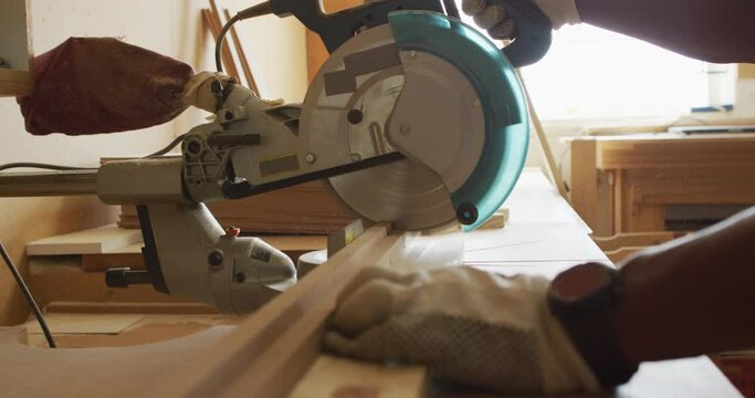 Close up of african american male carpenter hand's cutting wooden plank with electric chop saw