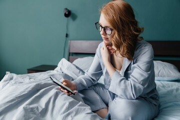 young red-haired caucasian woman in glasses and pajamas sits on the bed in the morning and checks...