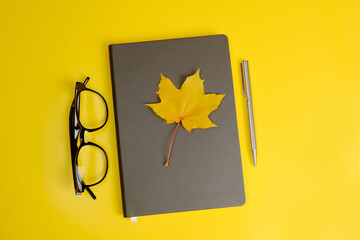 On a gray notebook there is a yellow maple leaf, glasses and a pen on a yellow background.Teacher's day concept, things for businessmen, education,autumn planning,autumn discounts on optics