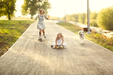 Foto auf Leinwand Cute red-haired girl mom hipster with tattoo rides skateboard with daughter child and dog of Australian Shepherd breed sidewalk in park, warm summer evening and family time and hobbies © natalialeb