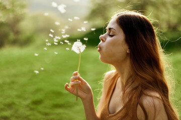 Woman with a dandelion flower in her hands smiling and blowing on it against a background of summer greenery and sunlight