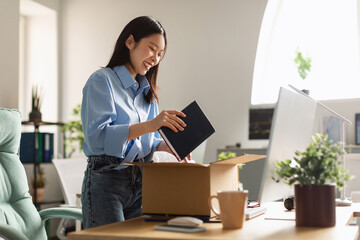 Smiling Asian woman unpacking box at new workplace - obrazy, fototapety, plakaty
