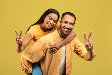 Handsome young African American man giving piggyback ride to his girlfriend, showing peace gesture on yellow background