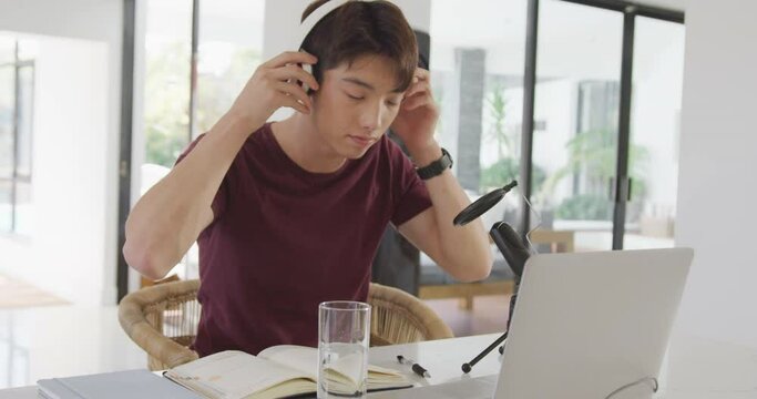 Asian boy wearing headphones speaking on professional microphone to record audio podcast at home