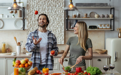 Glad caucasian millennial male with stubble juggles tomatoes, blonde lady prepares salad