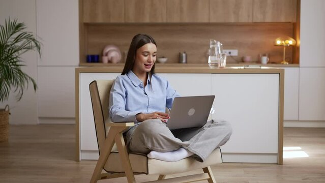 Young woman is calling online in front of laptop and sitting in chair at home interior spbas. 4k Beautiful American female with bionic prosthesis looks at display and talks with smile, holds device on