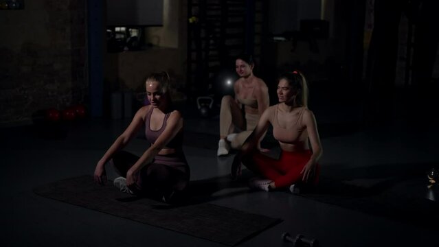 Wide shot joyful young women start training in gym looking away sitting down on exercise mats. Positive cheerful Caucasian slim sportswomen working out in darkness indoors. Slow motion