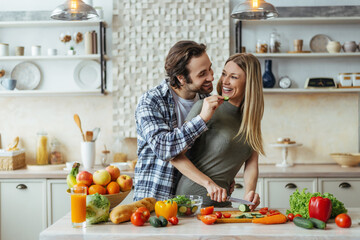 Happy caucasian millennial husband with stubble feeds blonde wife, couple have fun, lady prepare...