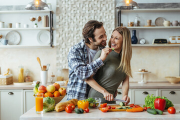 Happy caucasian millennial husband with stubble feeds blonde wife, couple have fun, lady prepare salad