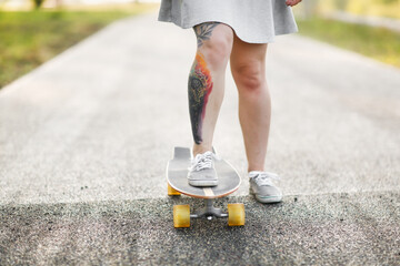 Beautiful female legs with tattoo on skateboard close-up in summer on path in park, generation z and sports walks