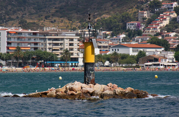 Small lighthouse at the port entrance of Roses city with beach and hotels in the background,...