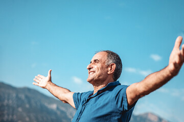 An aged happy smiling person standing with outstretched arms on mountains and blue sky background. - Powered by Adobe