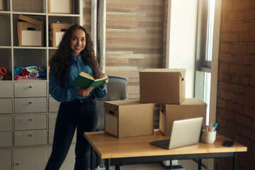 A young curly-haired logistics employee packs goods, collects parcels, puts things in a cardboard box, prepares a parcel for shipment, transports, sits on a windowsill and drinks a report in a book