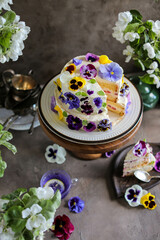 Obraz na płótnie Canvas Very beautiful two-tier cake with curd cream decorated with edible viola flowers. Cake with spring flowers on a high wooden stand.