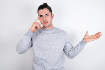 Young handsome dark haired man wearing fitted T-shirt over white wall confused and annoyed with open palm showing copy space and pointing finger to forehead. Think about it.