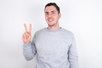 Fototapeta premium Young handsome dark haired man wearing fitted T-shirt over white wall showing and pointing up with fingers number two while smiling confident and happy.
