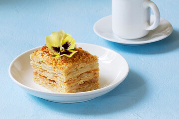 A slice of Napoleon cake, decorated with a viola flower, served with a cup of coffee or tea.