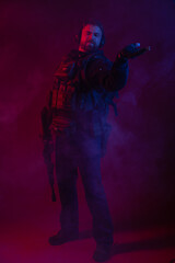 Fototapeta na wymiar soldier in full gear with weapons. a man in headphones, body armor, with a backpack and a belt. red background. colored, blue-red light. smoke around the military. explosion, chemical attack
