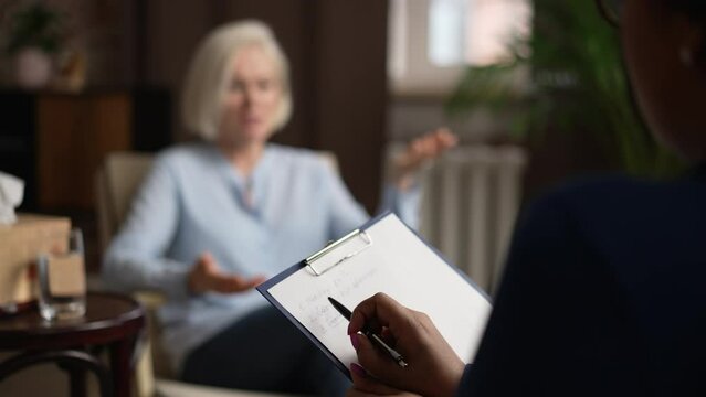 Ederly female patient is consulting with psychologist and sitting in modern office spbas. 4k Beautiful mature woman tells story and gestures with hands, experienced psychoanalyst listens to her and