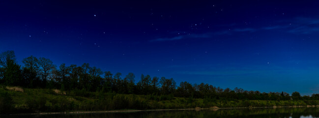 Starry night sky by a lake with trees in the background.