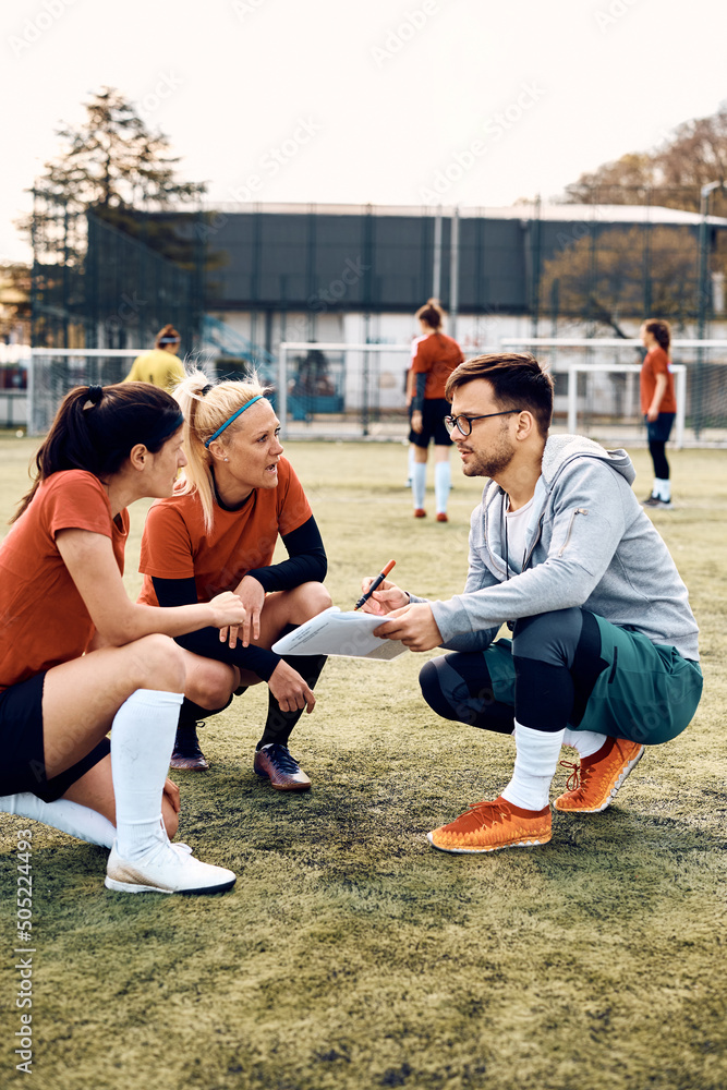 Wall mural Young coach and female soccer players analyzing game plan on playing field.