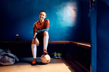 Portrait of happy female soccer player with ball in locker room looking at camera.