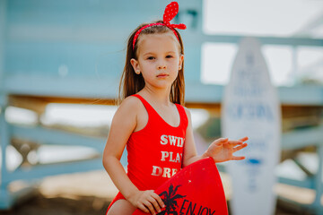 Pretty little girl in red bikini posing with small surfboard like a model on the beach against blue...