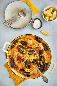 Classic dish of Spain, seafood paella in pan on light blue background top view. Spanish paella with shrimps, clamps, mussels, green peas and fresh lemon wedges from above