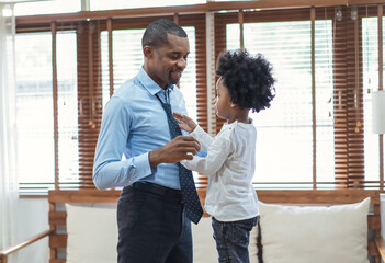 Happy African American businessman enjoying while little son is helping father tie necktie in...