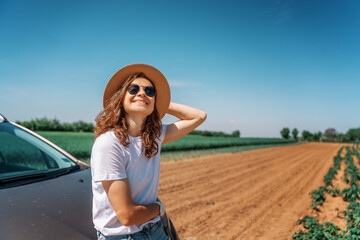 Happy cheerful young woman in hat and sunglasses traveling by car in the countryside on a summer day