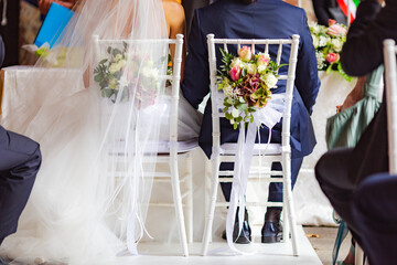Back view bride and groom sit on stylish retro chairs together in church wedding ceremony with bouquet