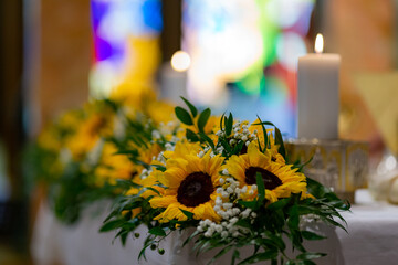 Flower decoration composed of sunflowers on the altar of a catholic church. Some burning candles and a colourful glass window in the background.