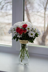 A bouquet of white chrysanthemums with a bush rose, coral in color, stands in a transparent vase with gold rims on the window. In front of the vase is a card in the form of a pink heart.