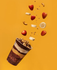 Acai cup, layers of granola, ground peanuts, and pieces of fruits and nuts flying in the air, yellow background.