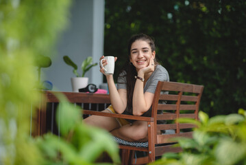 Pensive and thoughtful young smiling woman relaxing and breathing fresh air while sitting in balcony with nature view and drinking morning coffee and beverage