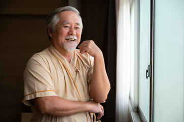 Cheerful and happy senior male patient standing near window in hospital room looking out and thinking while laughing