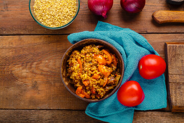 A plate of bulgur with vegetables and mushrooms next to onions and tomatoes and a cup of bulgur on a wooden table.