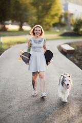 Naklejka na ściany i meble Cute red-haired hipster woman girl with tattoos in dress rides skateboard with an Australian Shepherd dog on sidewalk in park, warm summer evening