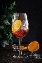 Shining cocktail Aperol syringe in a wine glass with ice and a slice of orange on a dark background with tree leaves